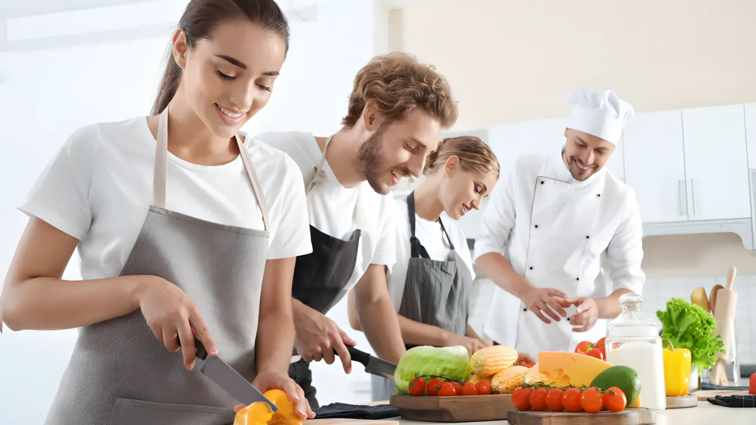 A group of four people in aprons preparing food in a kitchen. The woman in the center is holding a knife and chopping vegetables on a chopping board, while the man to her right is wearing a chef's hat. On the table in front of them are a variety of vegetables, a bottle, and other objects. In the background, there are cupboards and a wall.