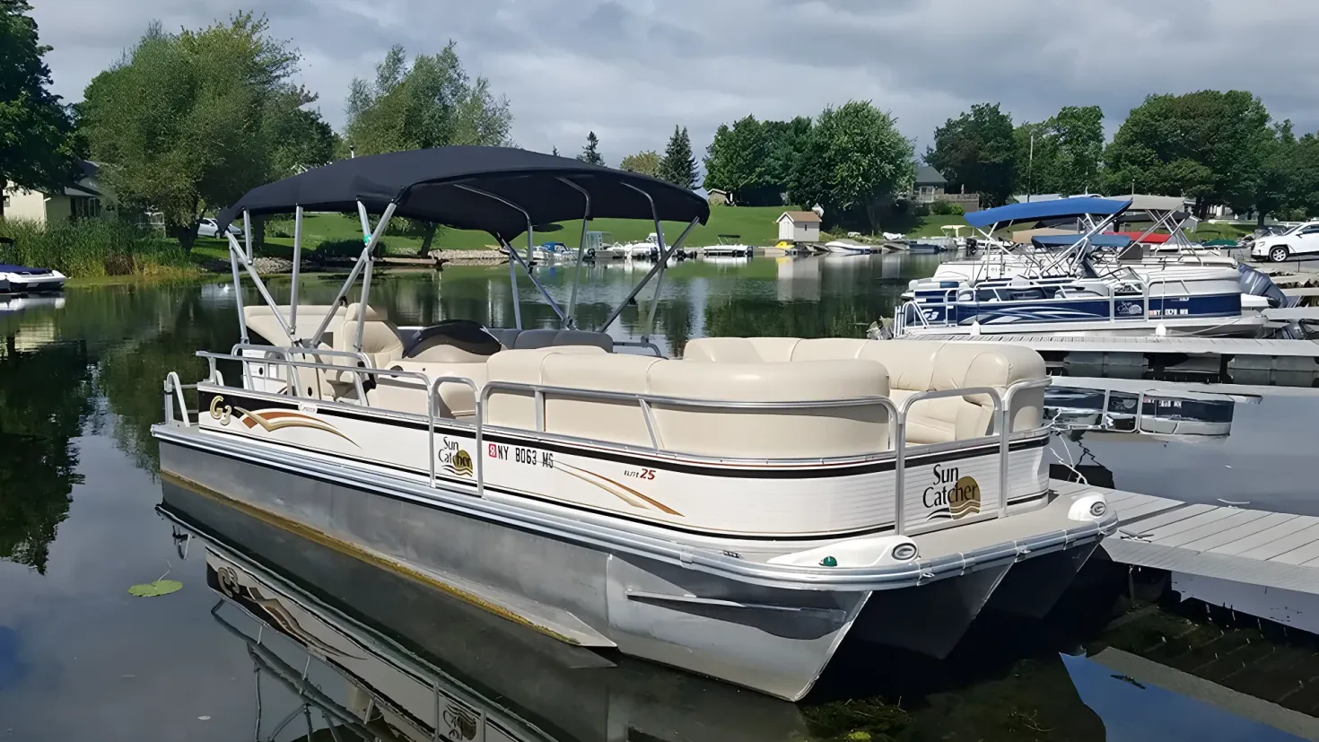 A pontoon boat docked at a dock with other boats in the background. On the right side of the image there is a wooden bridge, and on the left side there are houses and trees. The sky is visible at the top of the picture.