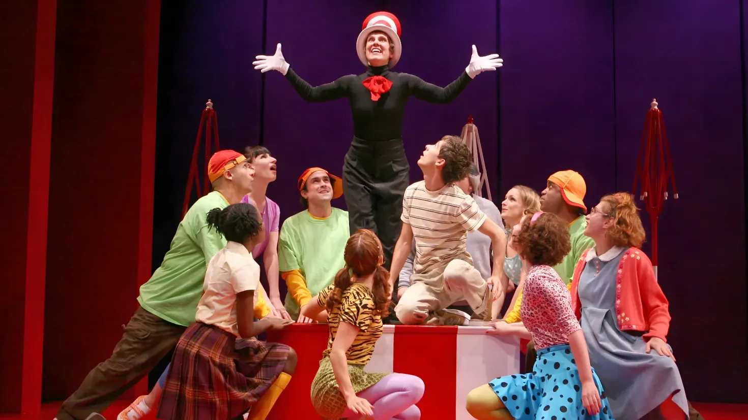 A group of people sitting on top of a stage, with a person standing in the center wearing a costume. The stage is decorated with curtains in the background, creating a vibrant atmosphere.