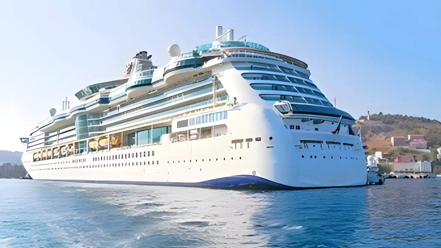 A large white cruise ship docked in the water near a dock, surrounded by buildings, trees, hills and a clear blue sky.