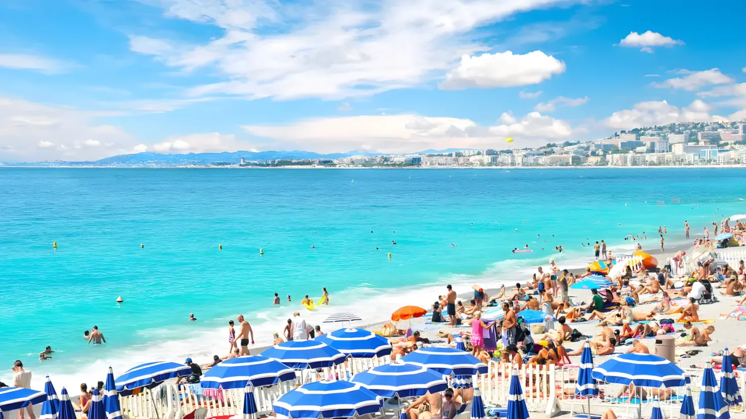 A crowded beach with many people enjoying the sun and sand. There are umbrellas providing shade from the sun, and a railing along the shoreline. In the background, there are buildings, hills, and clouds in the sky.