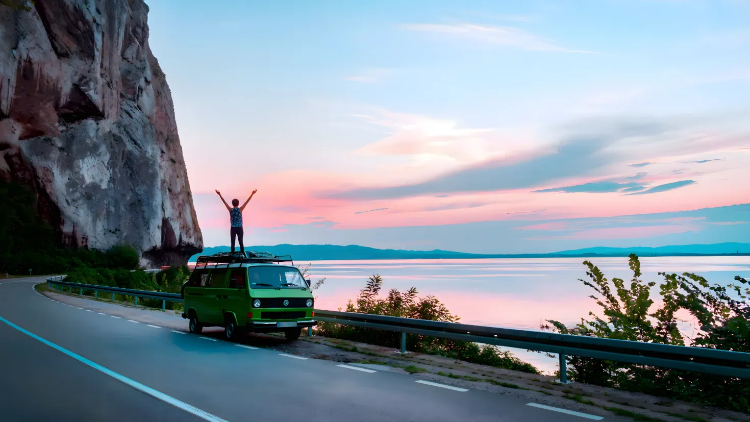 A person standing on top of a green van on the side of a road, surrounded by a railing, plants, and a large rock. In the background, there is a body of water, hills, and clouds in the sky.