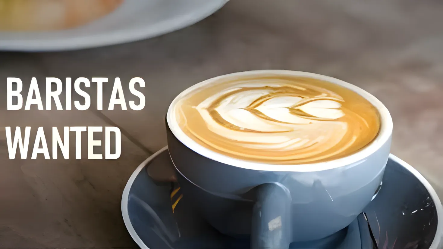 A cup of coffee with the words "Baristas Wanted" on it, sitting on a saucer with a spoon beside it. On the left side of the image, there is a plate with a food item on it.