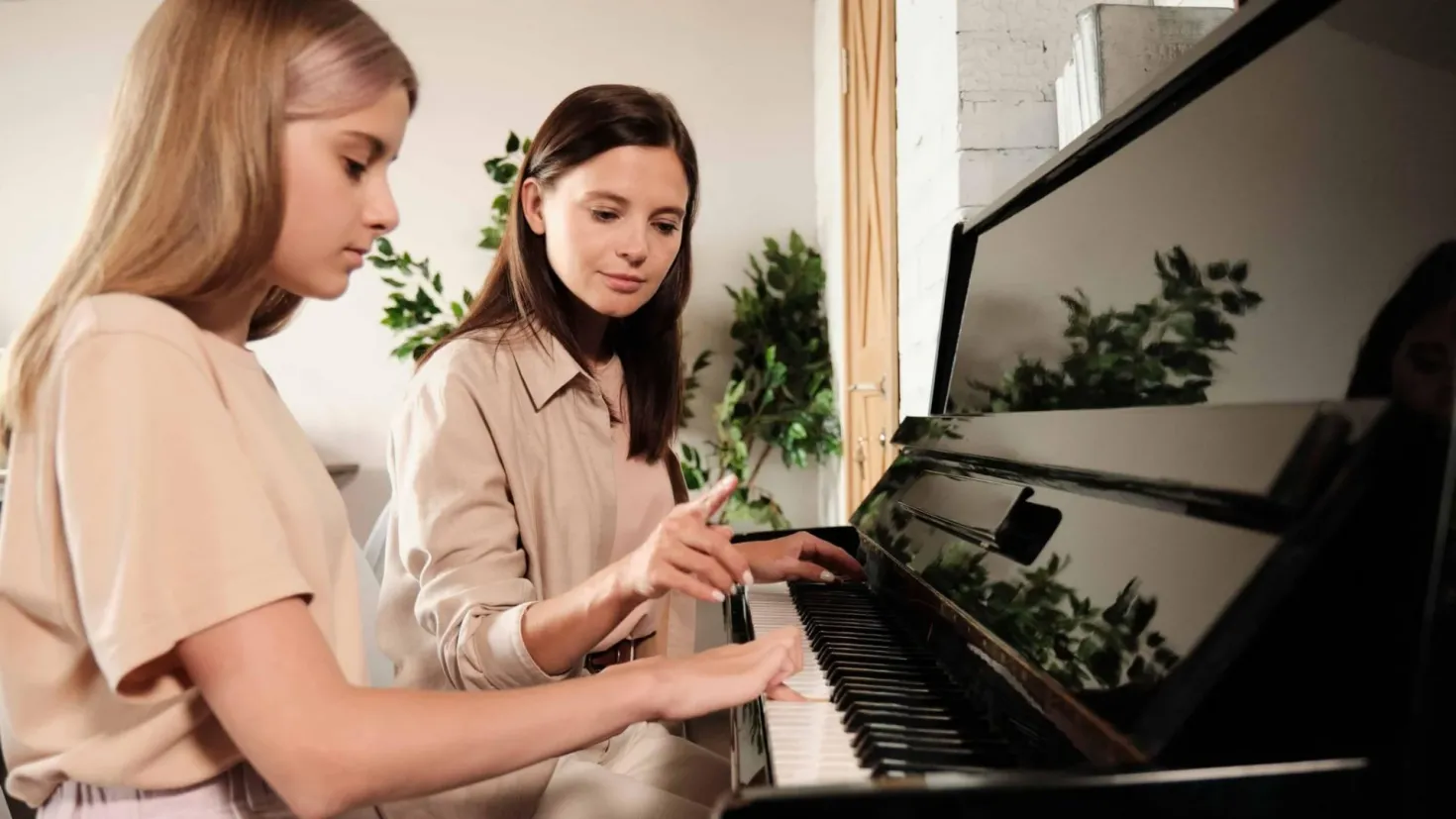 Two women sitting at a piano in a living room. The woman on the left is wearing a cream-colored dress and is playing the piano, while the other woman is listening intently. In the background, there are a few plants and a white wall.