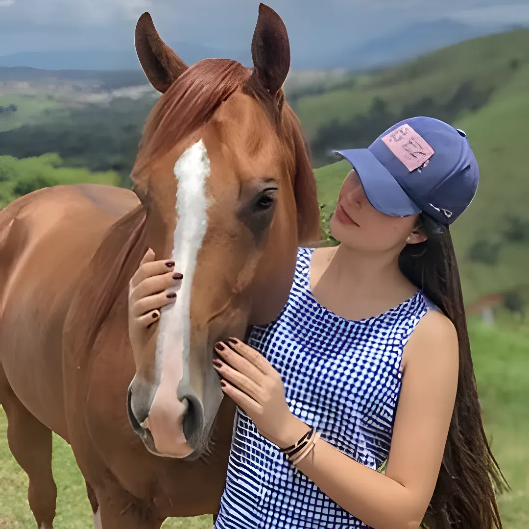 A woman wearing a blue and white dress and a blue cap, embracing a brown horse in a field surrounded by trees and mountains in the background.