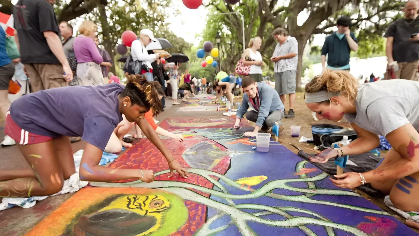 A group of people painting on the ground in front of a crowd of people, some of whom are holding umbrellas and handbags. There are also bottles, glasses, and other objects scattered around the area. In the background, there are trees and balloons, and the sky is visible above them.
