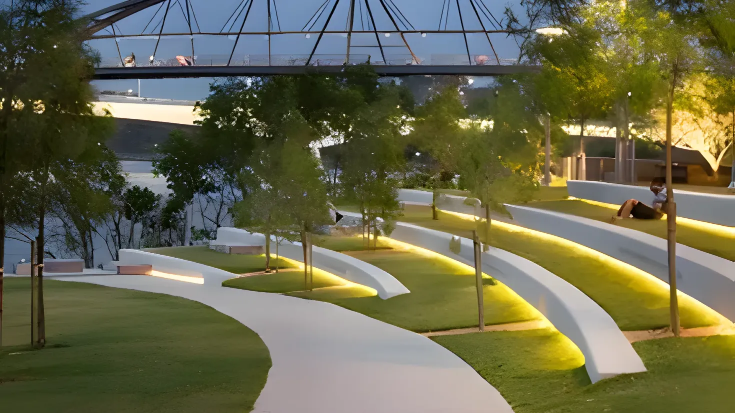A park with a walkway in the middle of it, surrounded by lush green grass, trees, and light poles. There is a person sitting on the grass, and in the background there is a bridge with rods and lights, as well as a body of water. The sky is visible at the top of the image.