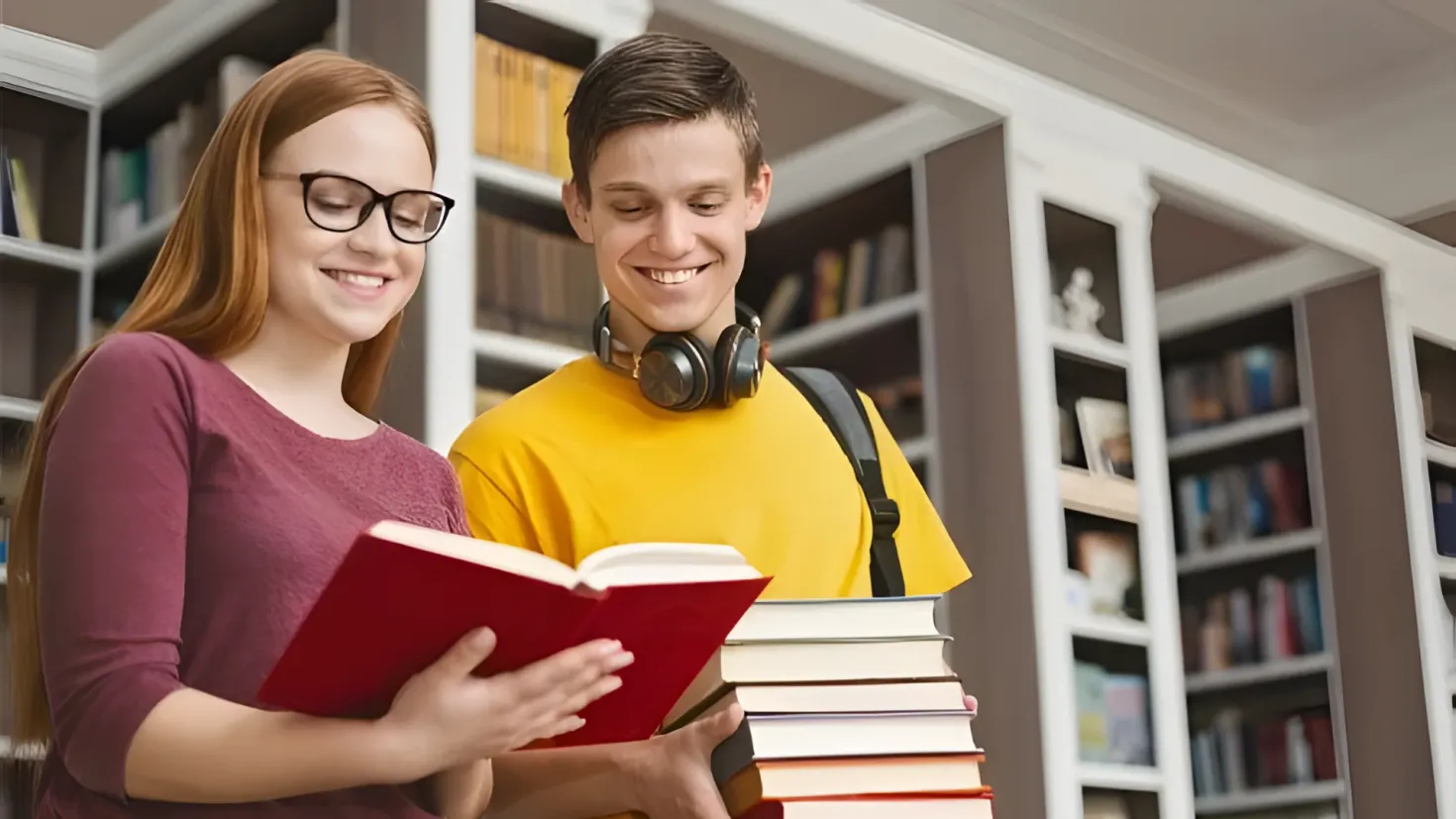 A man and woman standing in a library, both of them smiling and holding books. The man is wearing a yellow t-shirt and headphones, while the woman is wearing spectacles. In the background, there are bookshelves filled with books.