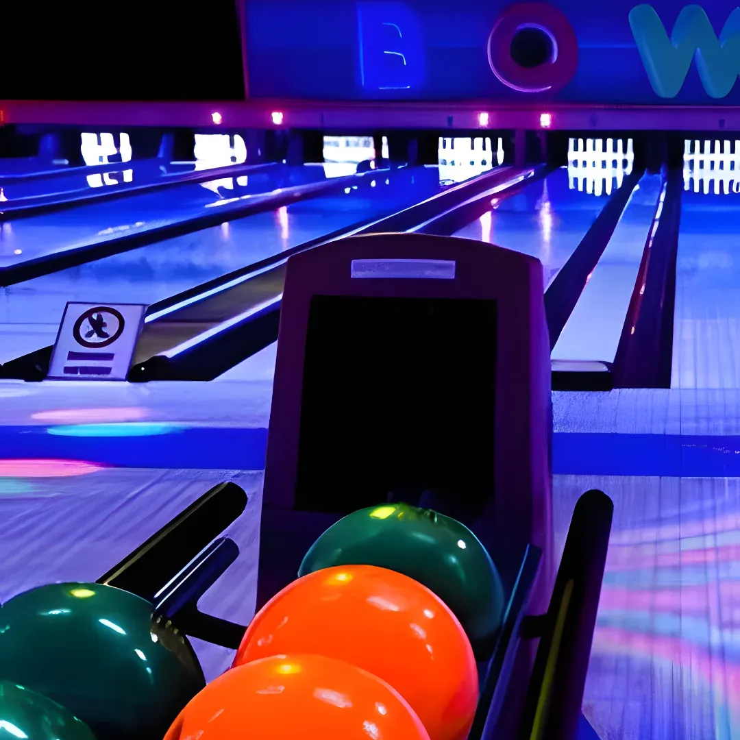 A bowling alley with colorful bowling balls lined up in the lanes, illuminated by lights and a board with text in the background.