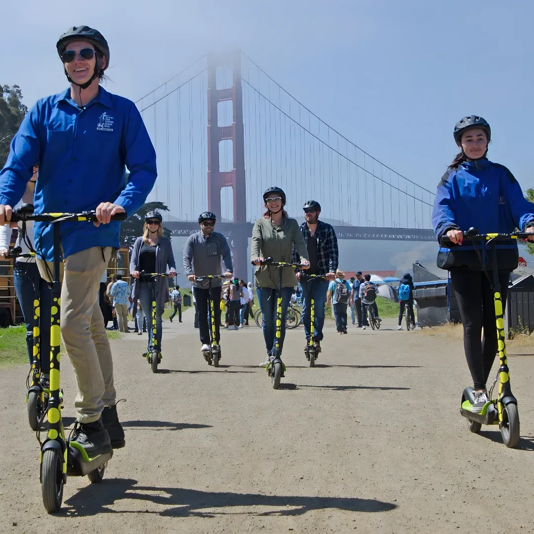 A group of people riding electric scooters in front of the iconic Golden Gate Bridge in San Francisco, California. The people are wearing helmets and goggles, and the ground is covered in grass. In the background, there are trees, sheds, and a clear blue sky.