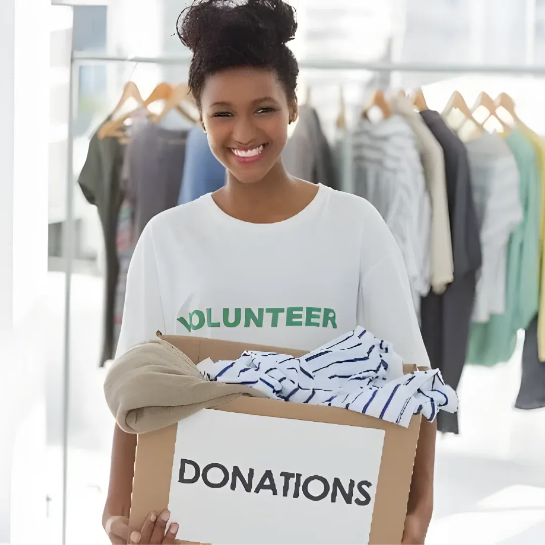A woman standing and smiling while holding a cardboard box with the word "Donations" written on it. Behind her, there are clothes hanging from hangers and a glass door.