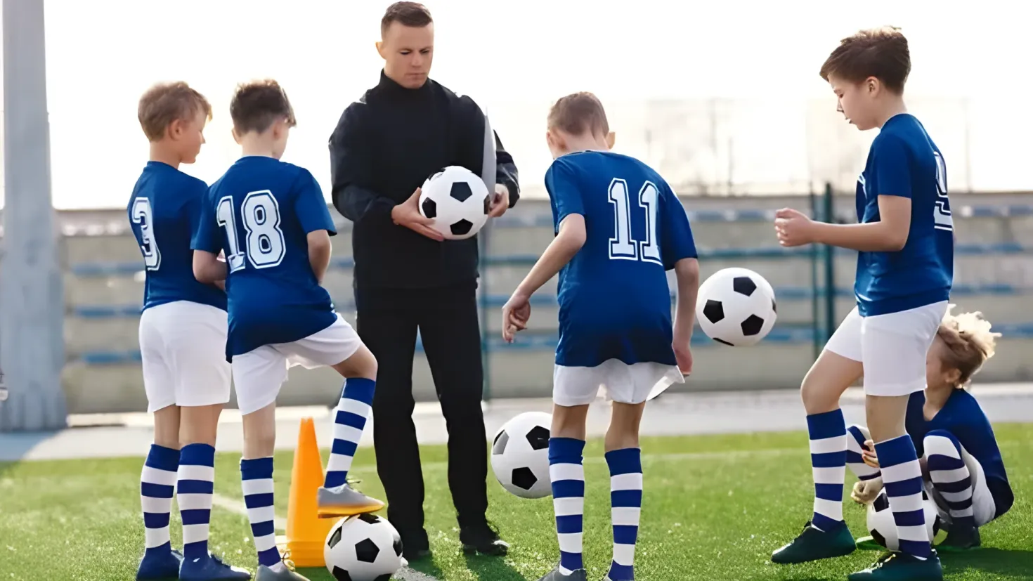 A group of young boys standing on top of a soccer field, with a man standing in the center holding a ball. There are several soccer balls scattered around them, and a traffic cone in the foreground. In the background, there are railings and a clear blue sky.
