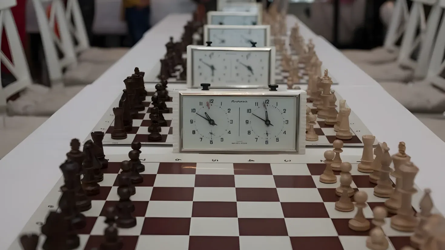 A chess board with chess pieces and a clock on it, surrounded by a few people in the background. The background is slightly blurred, giving the image a dreamy feel.