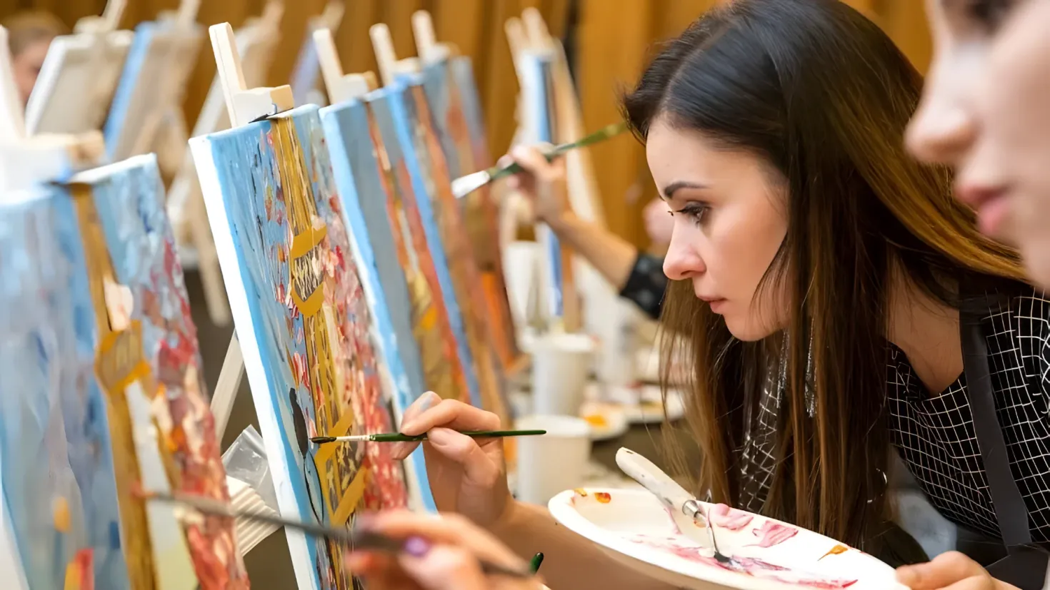 Two women painting on an easel in an art studio. They are both holding paint brushes and painting on the boards, with a plate of paint in front of them. In the background, there are other people and objects on the table.