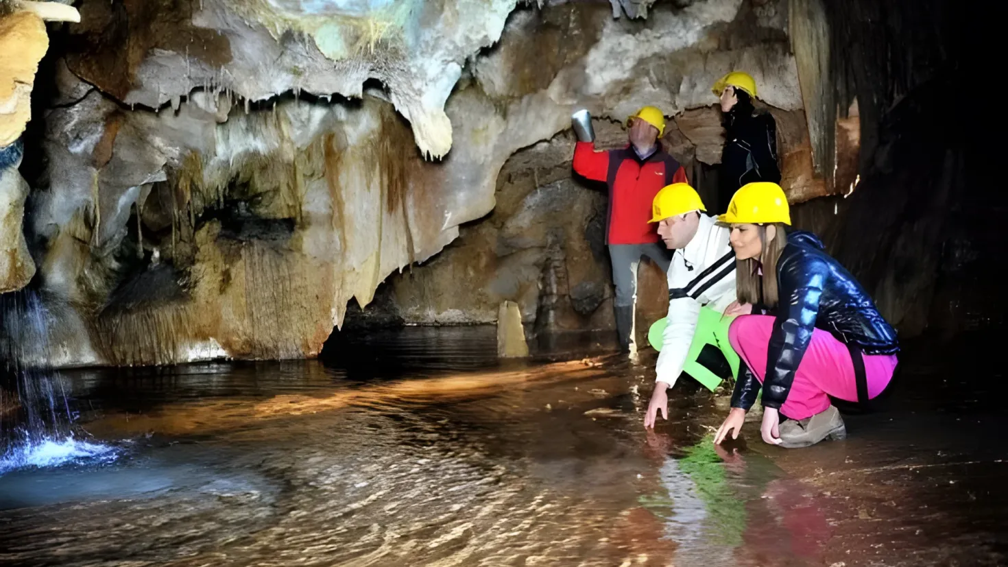 A group of people wearing yellow hard hats standing in a cave, with one person holding an object in their hand. The water is visible at the bottom of the image, and the cave walls are visible in the background.