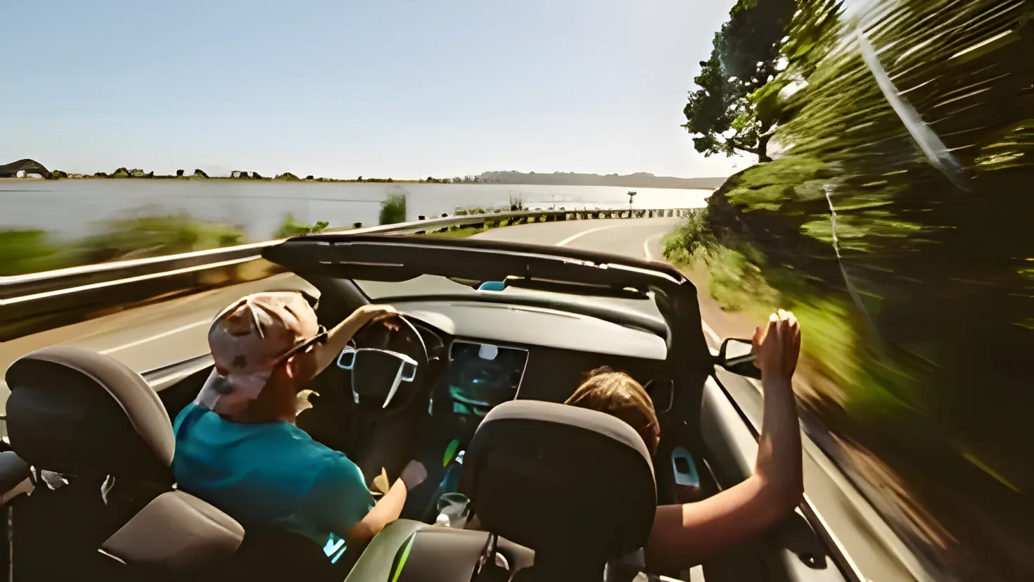 A man and woman driving a convertible car down a road next to a body of water, with trees on the right side of the road and a clear blue sky in the background.