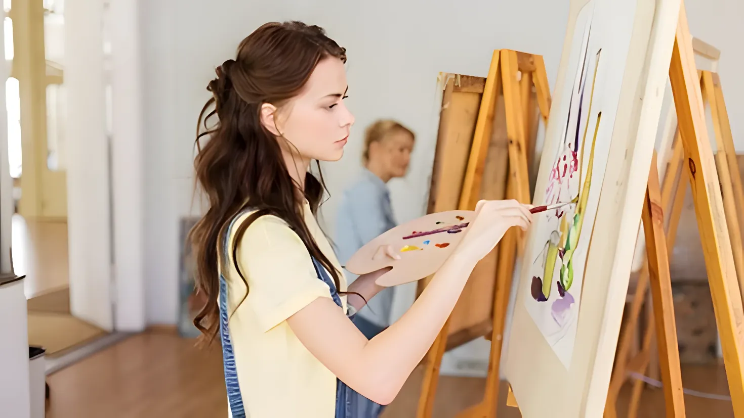 A woman painting on an easel in an art studio. She is holding a brush and painting on the board, while a man stands in the background. On the left side of the image, there is a table with various objects on it, and a wall behind them.