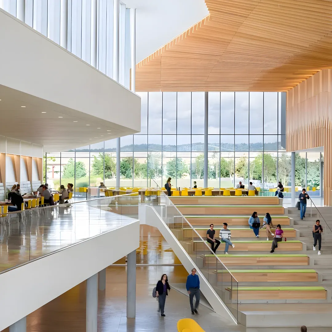 A large atrium filled with lots of people walking around, some of them sitting on chairs and some standing on the floor. There are stairs, railings, pillars, lights, and glass walls that offer a view of the trees, hills, and sky outside.