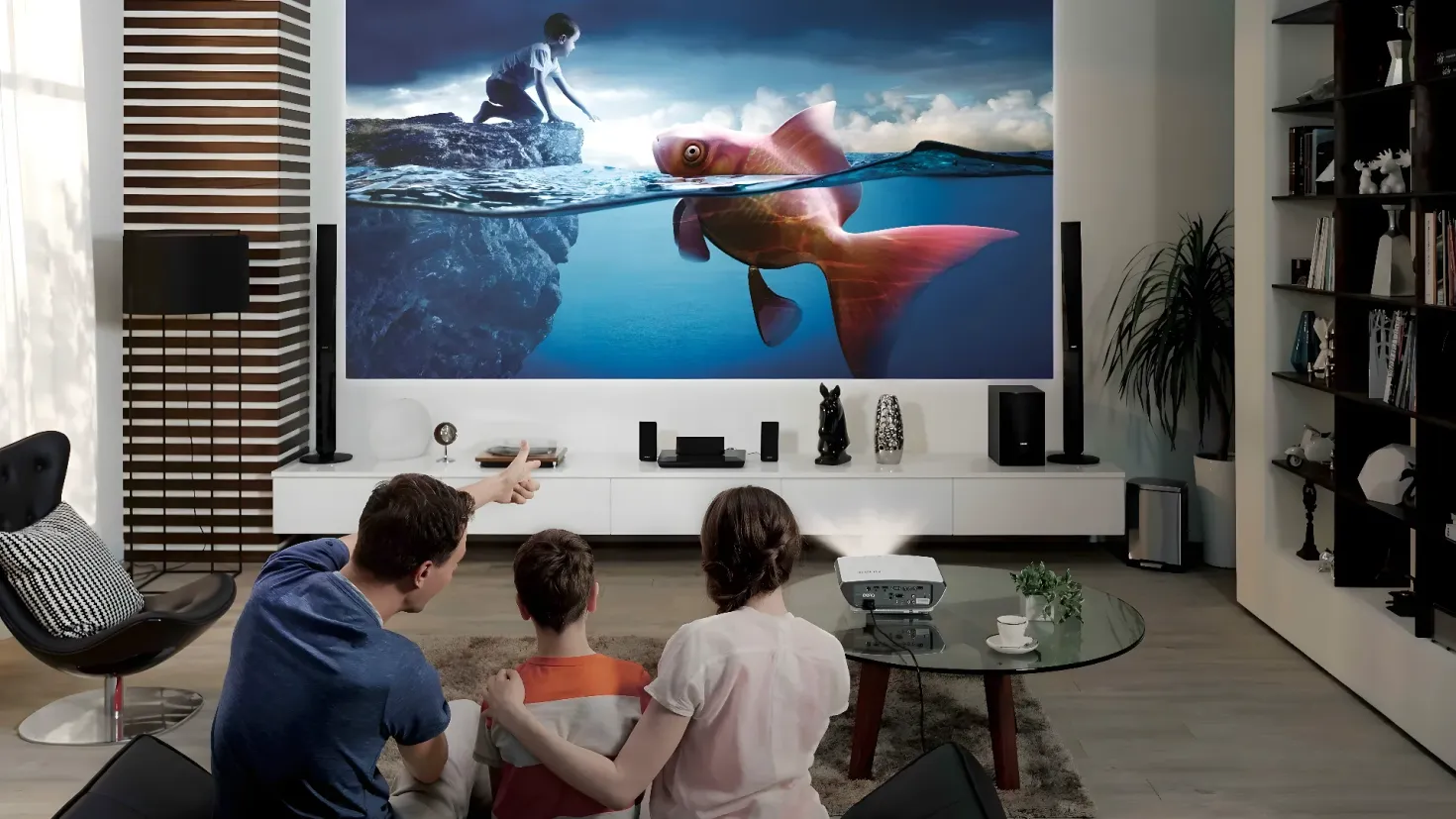 A group of people sitting around a table in front of a large flat screen TV. On the table there is a printer, a cup with saucer, a plant, and other objects. To the right of the table is a cupboard with books, toys, and a pot with a plant. In the background, there are speakers and other items on a platform, as well as a television on a stand. The floor is covered with a carpet.