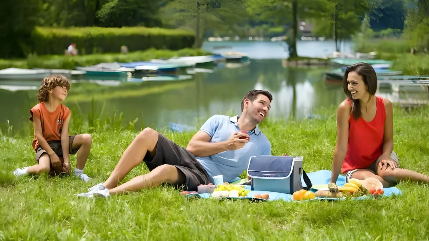 A family of four - a man, woman and two children - sitting on a blanket in the grass near a lake. The man is holding an object in his hand and there is a bag, fruits and other objects on the blanket. In the background, there are boats on the water, plants and trees.