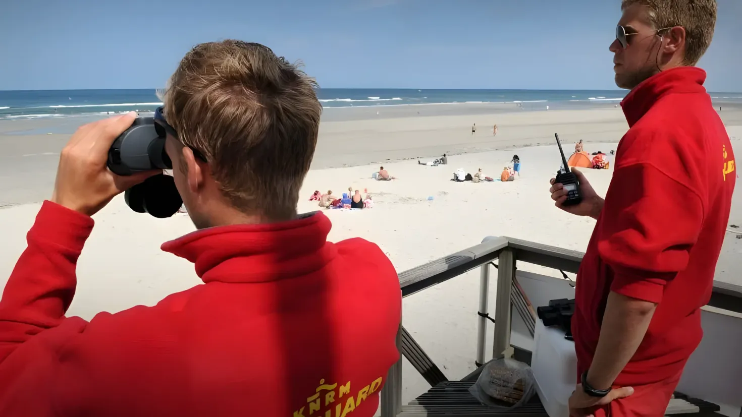 Two men in red shirts standing on a balcony overlooking the ocean. One of the men is holding a camera in his hand, while the other is wearing glasses and a bracelet. There are a few people sitting on the sand in the background, and the sky is visible above them.