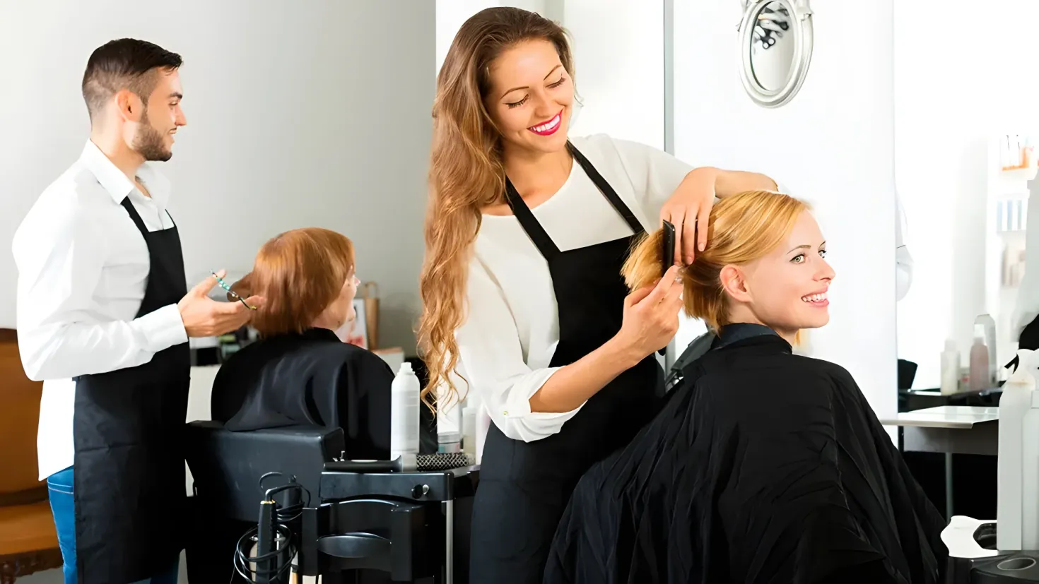A woman getting her hair cut in a salon. She is sitting in a chair with a smile on her face, while a hairdresser stands behind her, holding an object in her hand. On the right side of the image, there are bottles and other objects on the table, and in the background there is a mirror on the wall.