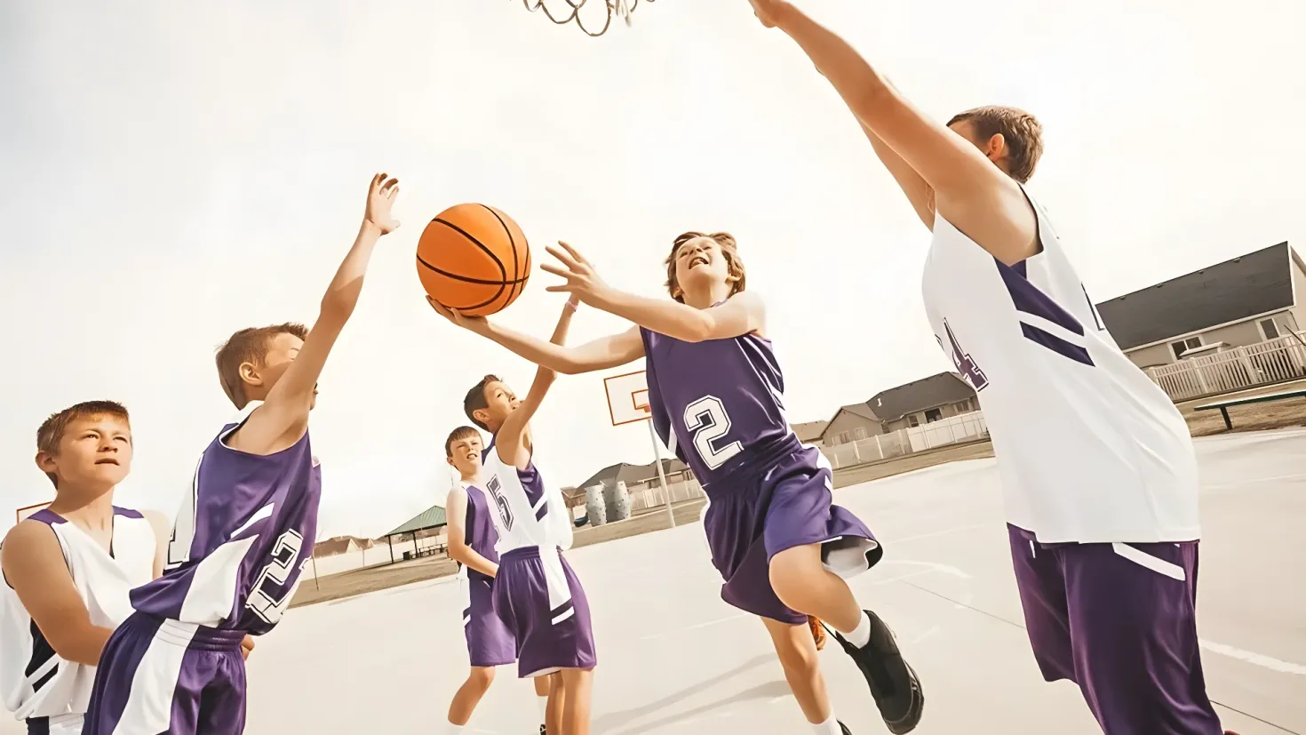 A group of young boys playing basketball on a court, with a basketball net at the top of the image. In the background, there are buildings, railings, and a clear blue sky.