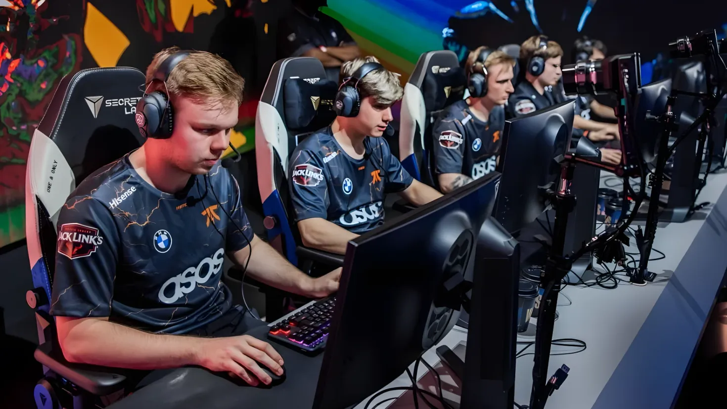 A group of young men sitting at a desk in front of a computer, wearing headphones and playing a game. On the desk there are monitors, keyboards, mouses and other objects, and in the background there is a wall with a painting.