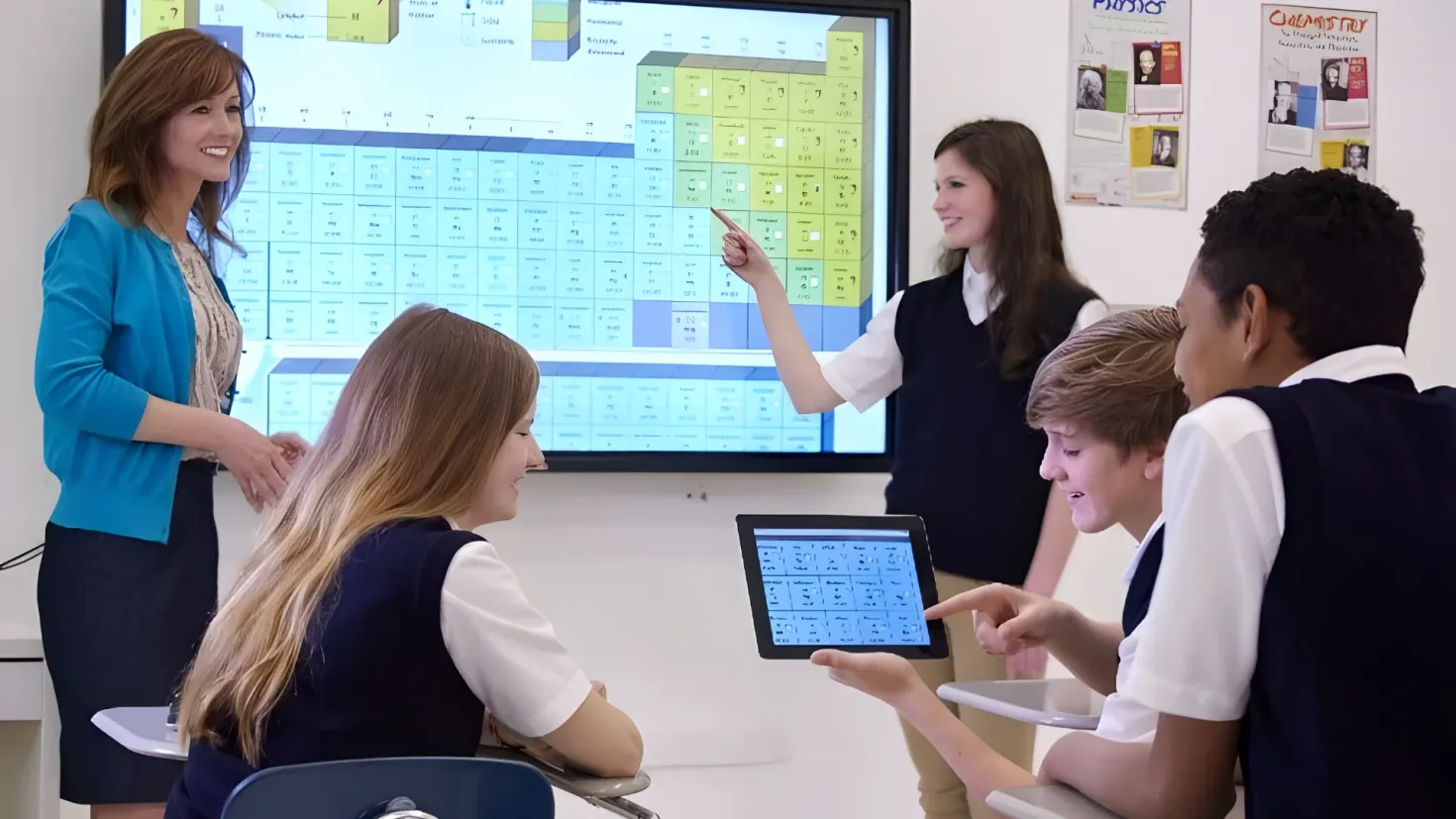 A group of students sitting at desks in front of a large screen, with a woman standing and smiling in the center. On the right side of the image, there are papers attached to the wall, and on the left side, there is a table with various objects on it.
