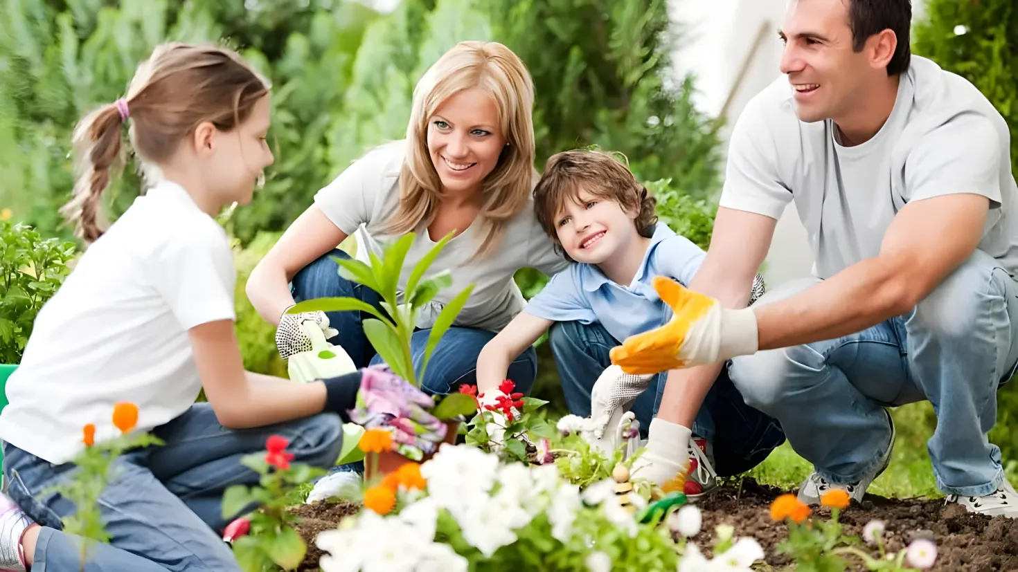 A family of four - a man, woman and two children - planting flowers in a garden. The man is wearing gloves and is holding a flower pot, while the woman and children are wearing white t-shirts and blue pants. The background of the image is filled with lush green plants and trees, and the ground is covered in soil.
