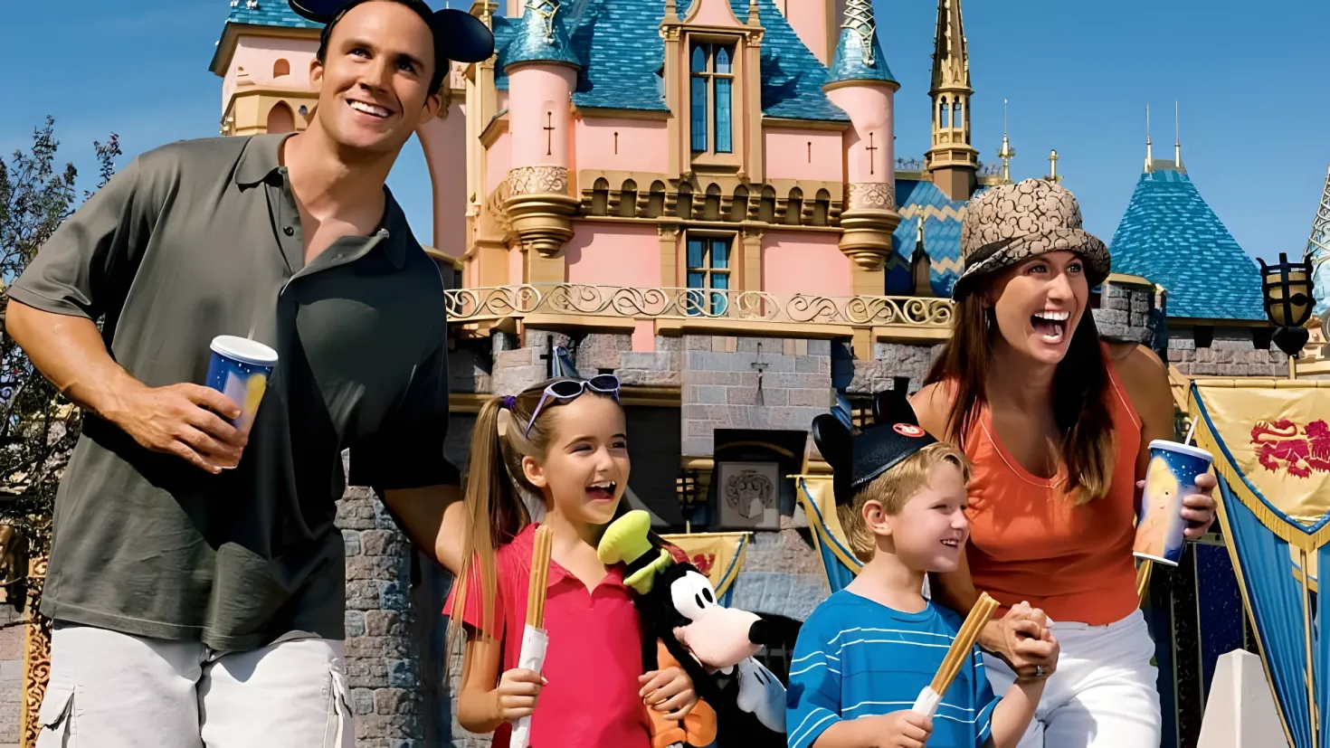 A family of four - a man, woman and two children - standing in front of Cinderella's Castle at Walt Disney World. The man is wearing a black t-shirt and white pants, while the woman and children are wearing colorful dresses and hats. The children are holding various objects in their hands, and the castle is surrounded by trees and a clear blue sky.