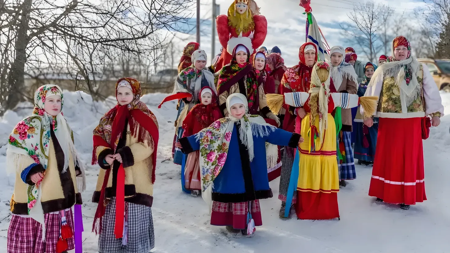 A group of people in traditional Russian clothing walking through the snow. They are wearing colorful dresses and some of them are holding flags. In the background, there are trees, poles, wires, vehicles, and a clear blue sky.
