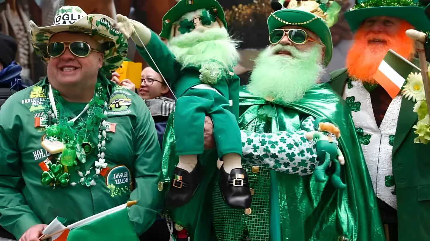 A group of people dressed in green and white outfits, including hats, glasses, and a garland, standing next to each other in front of a crowd of people. One person is holding an object in their hand, and in the background there are trees. It appears to be a St. Patrick's Day parade.