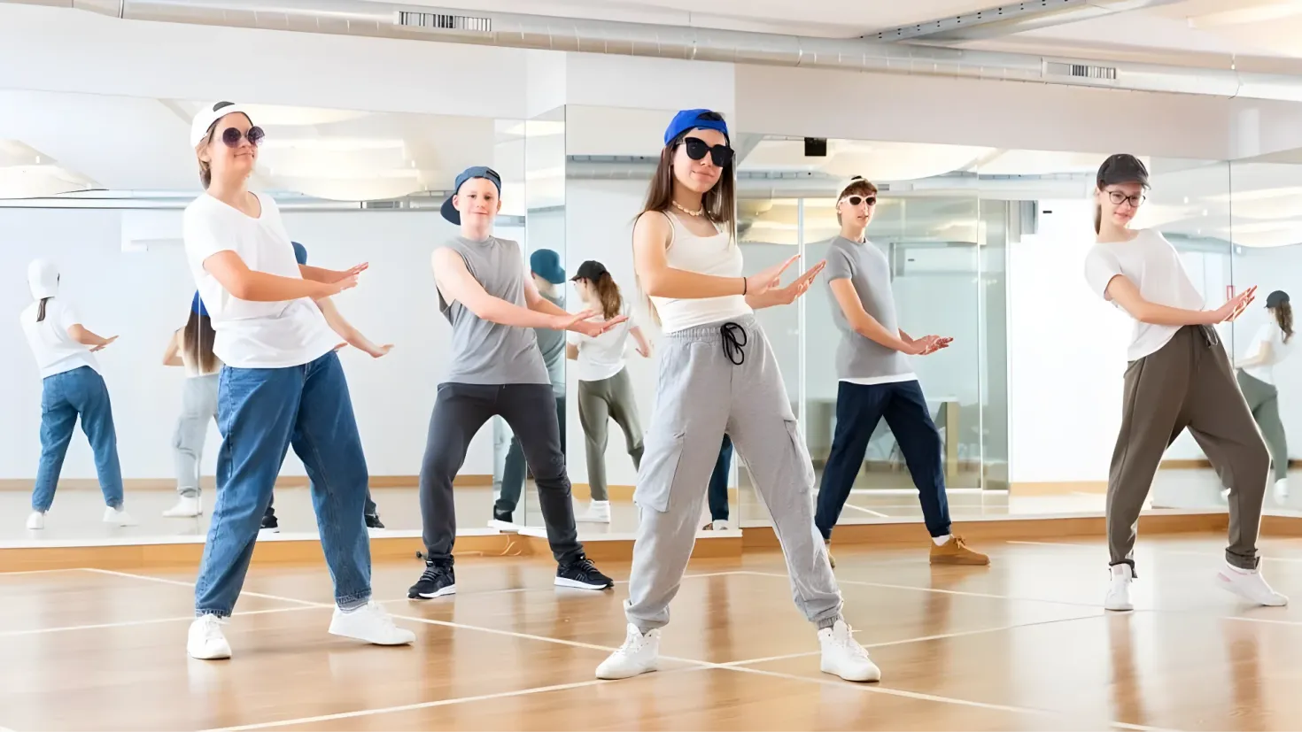 A group of people in a dance studio, wearing white t-shirts, blue caps, and glasses. They are dancing on the floor, with a wall in the background and lights on the ceiling.
