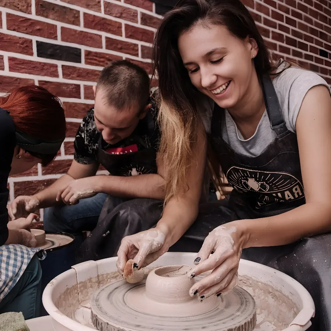 A group of people working on a pottery wheel in front of a brick wall. There is a woman and two men in the foreground, both of them smiling as they work on the pottery.