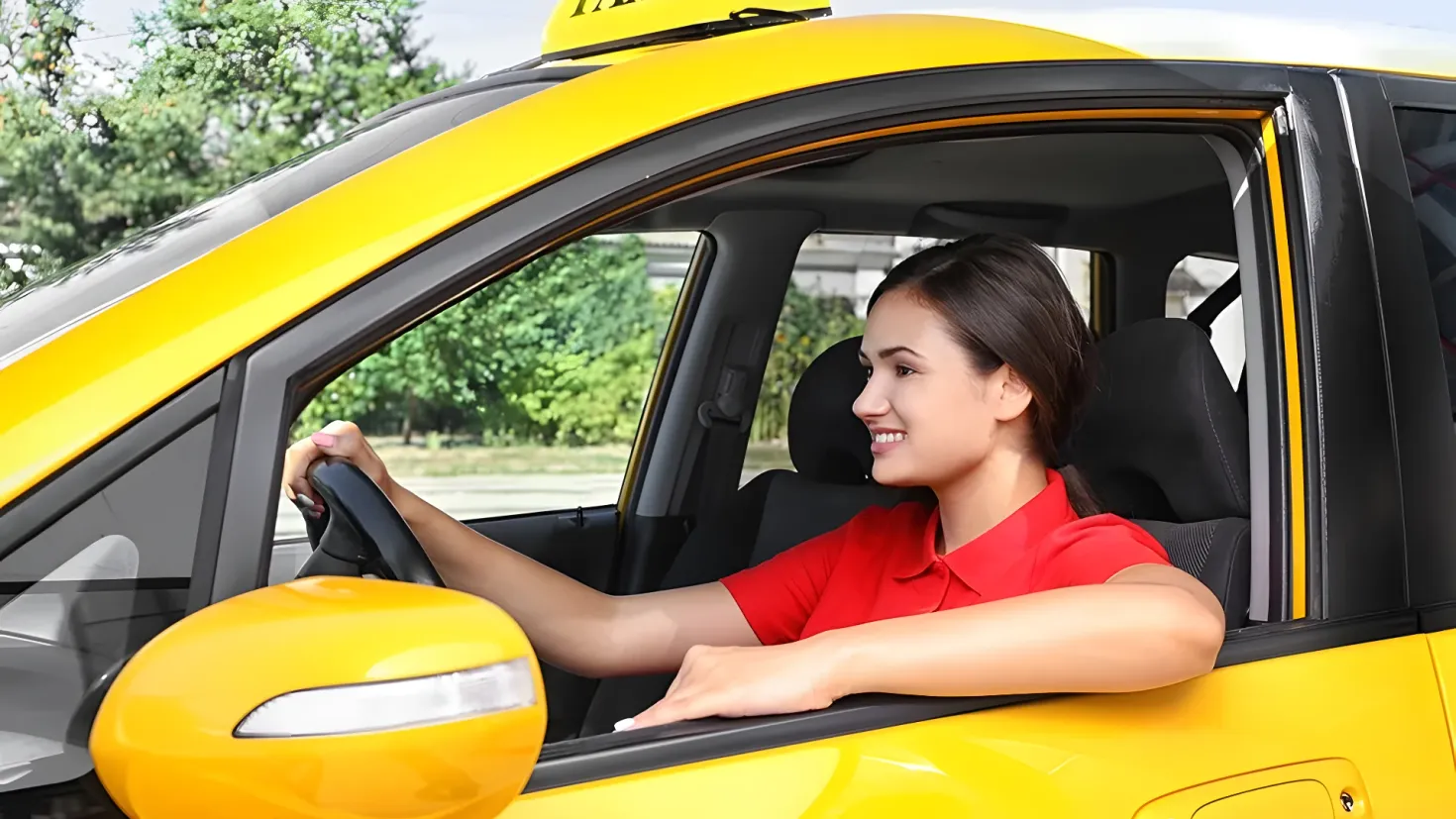 A woman driving a yellow taxi with a smile on her face. She is wearing a red t-shirt and the car is surrounded by trees and a clear blue sky.