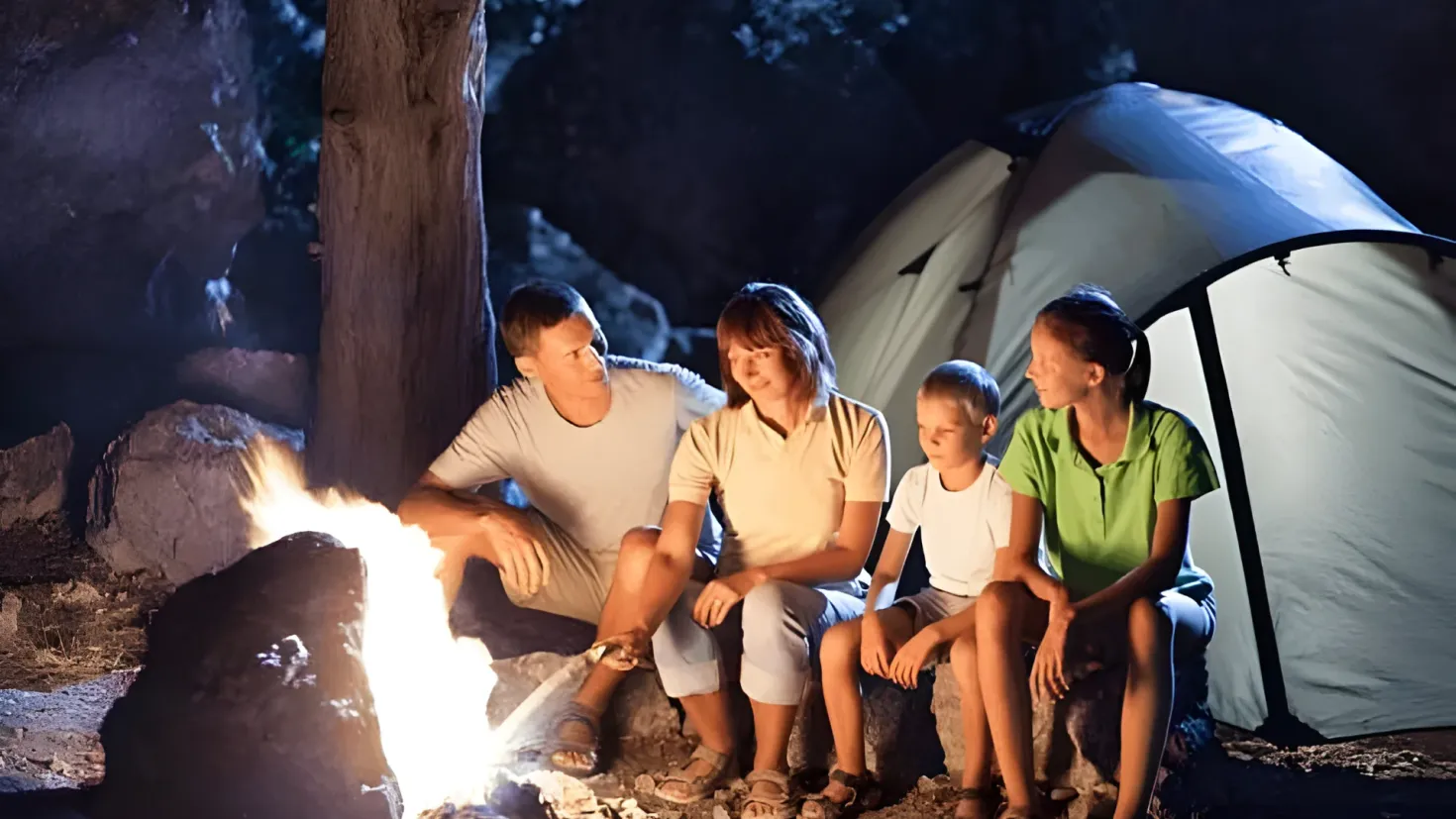 A family of four sitting around a campfire in the woods at night. The campfire is surrounded by rocks and a tree trunk, and there is a tent in the background. The family is illuminated by the warm glow of the fire, creating a cozy atmosphere.