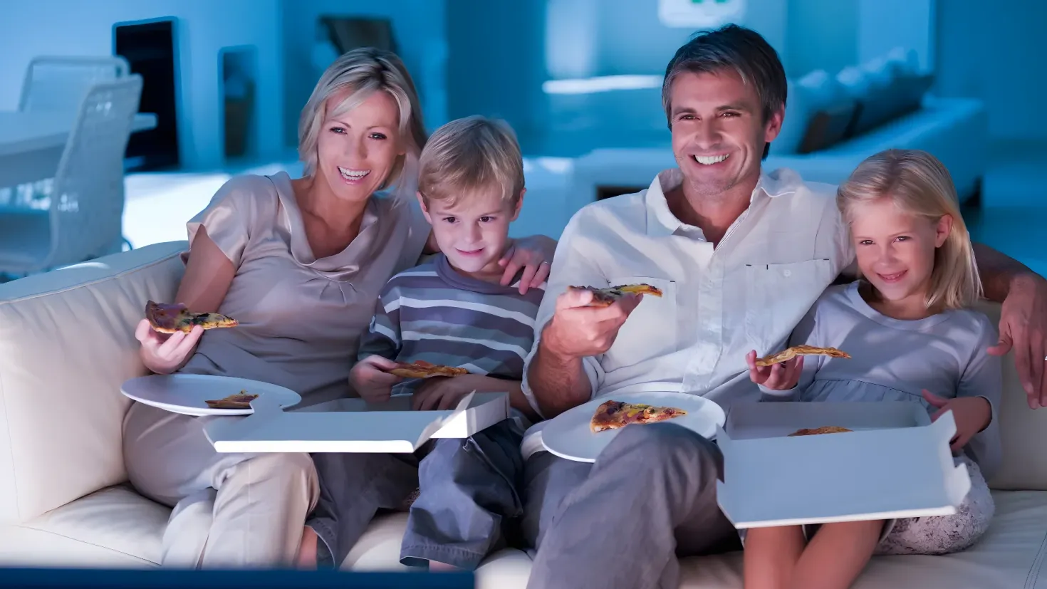 A family of four sitting on a couch, each holding a plate of pizza. In the background, there are chairs, a table, and a wall.