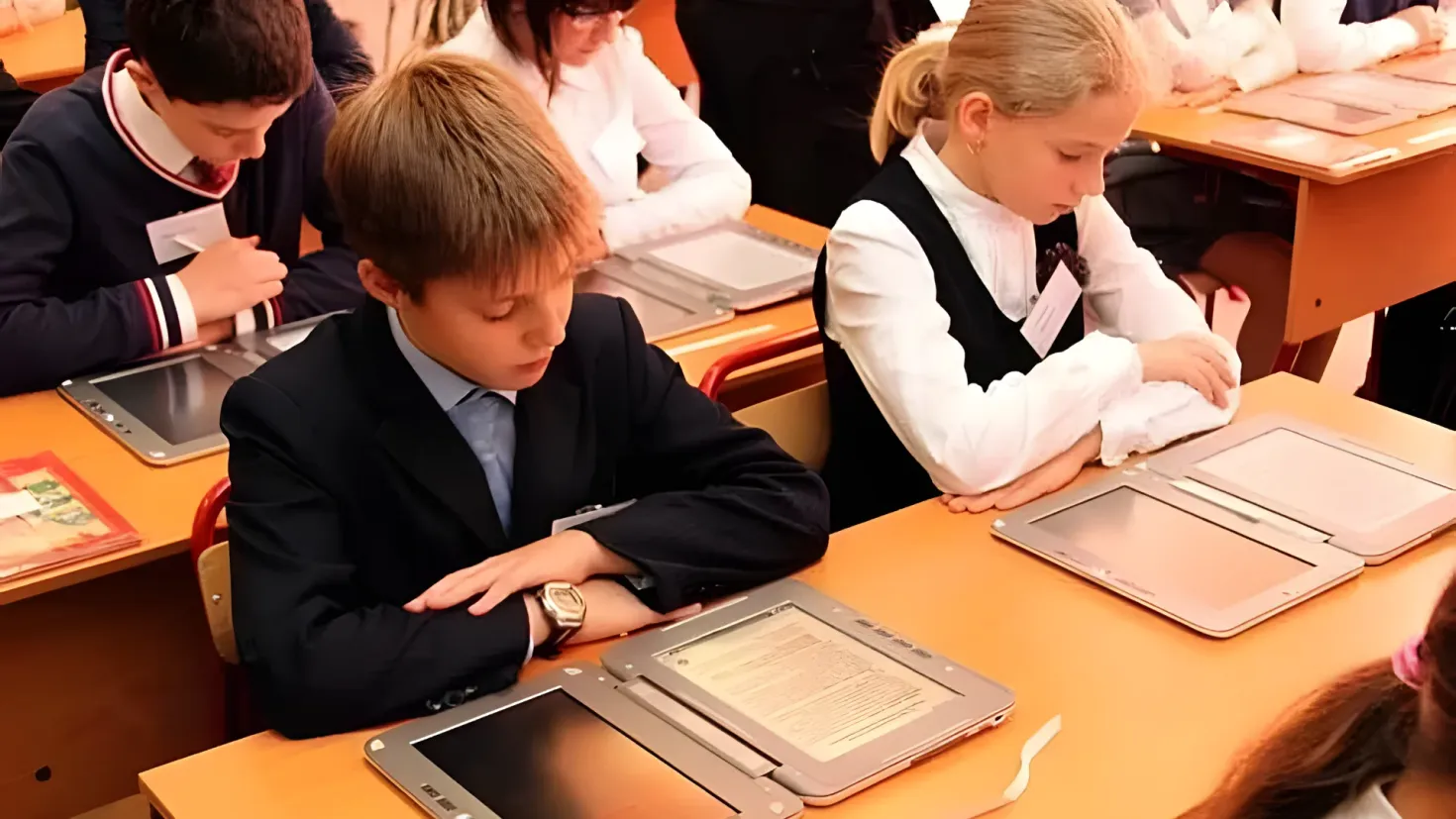 A group of children sitting at desks with iPads in front of them. There are books and other objects on the tables, and a person standing in the background.