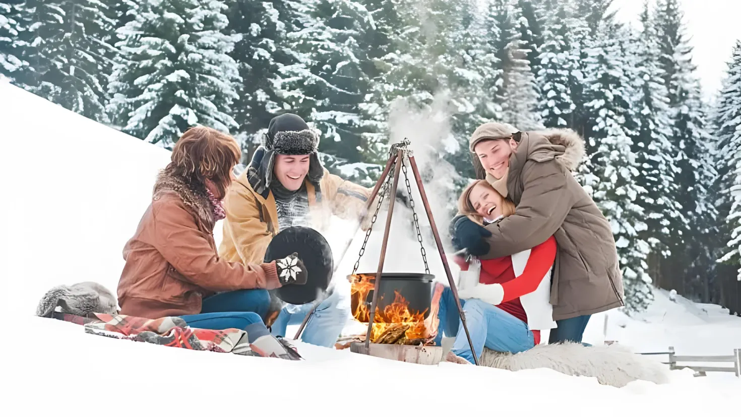 A group of four people sitting around a campfire in the snow. They are all wearing jackets and caps, and one of them is holding a black object. The campfire is surrounded by trees covered in snow, and there are various objects scattered around it.