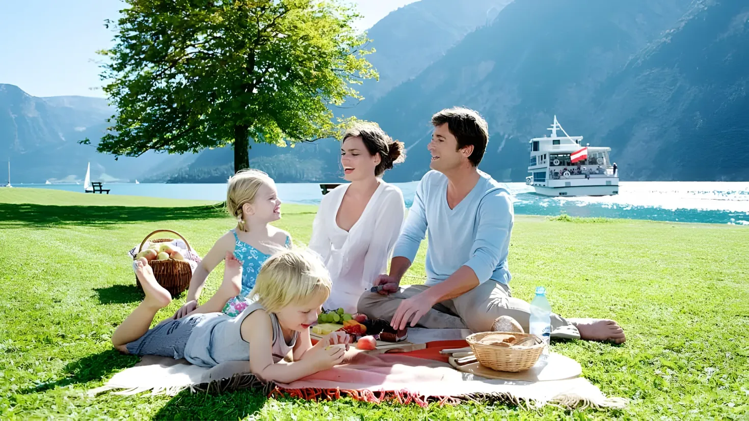 A family of four - a man, woman and two children - having a picnic on the grass in front of a lake. The lake is surrounded by trees and hills, and there is a boat on the water in the background. The sky is visible at the top of the image.