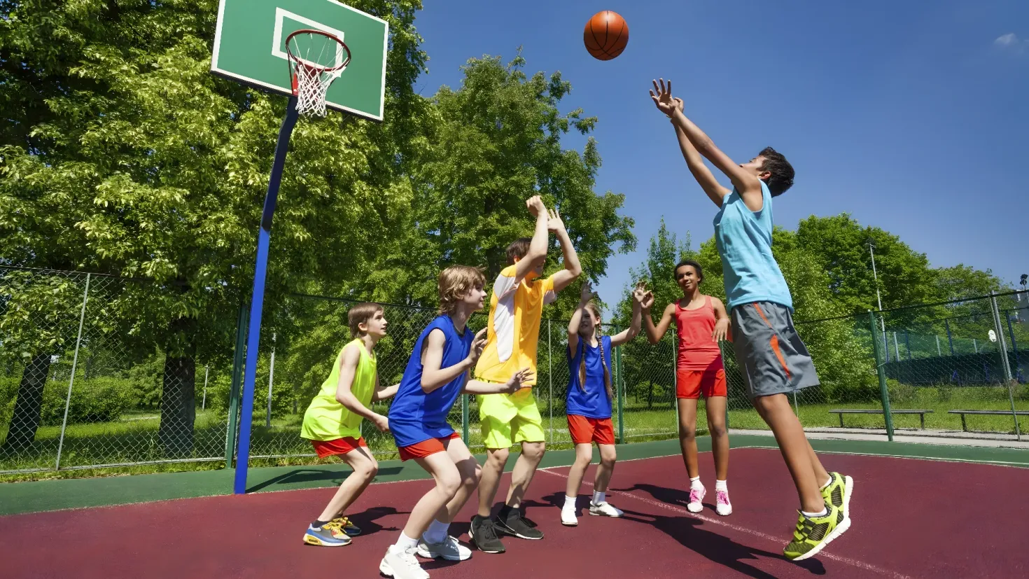 A group of young people playing basketball on a court surrounded by a fence, benches, grass, plants, trees, and a clear blue sky.