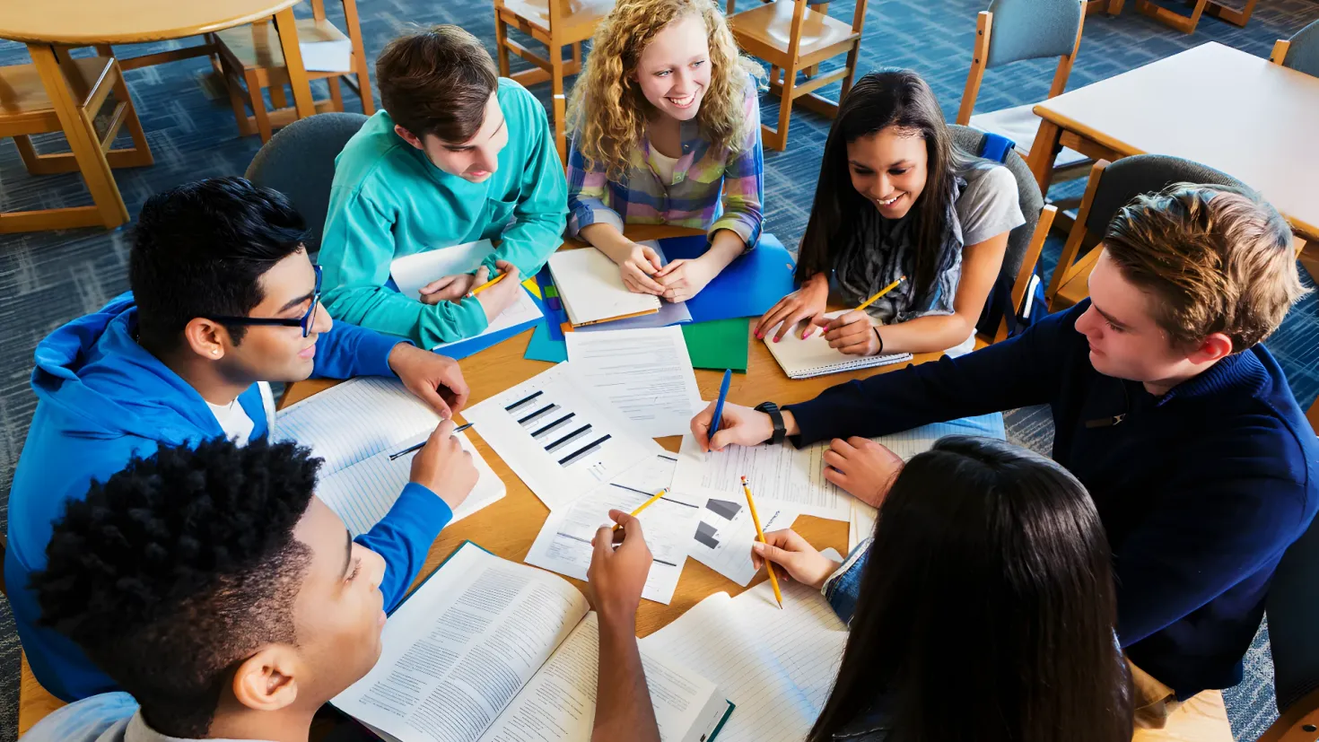 A group of students sitting around a table in a classroom, with books, papers, and pencils spread out in front of them. They are all smiling and appear to be engaged in a discussion.