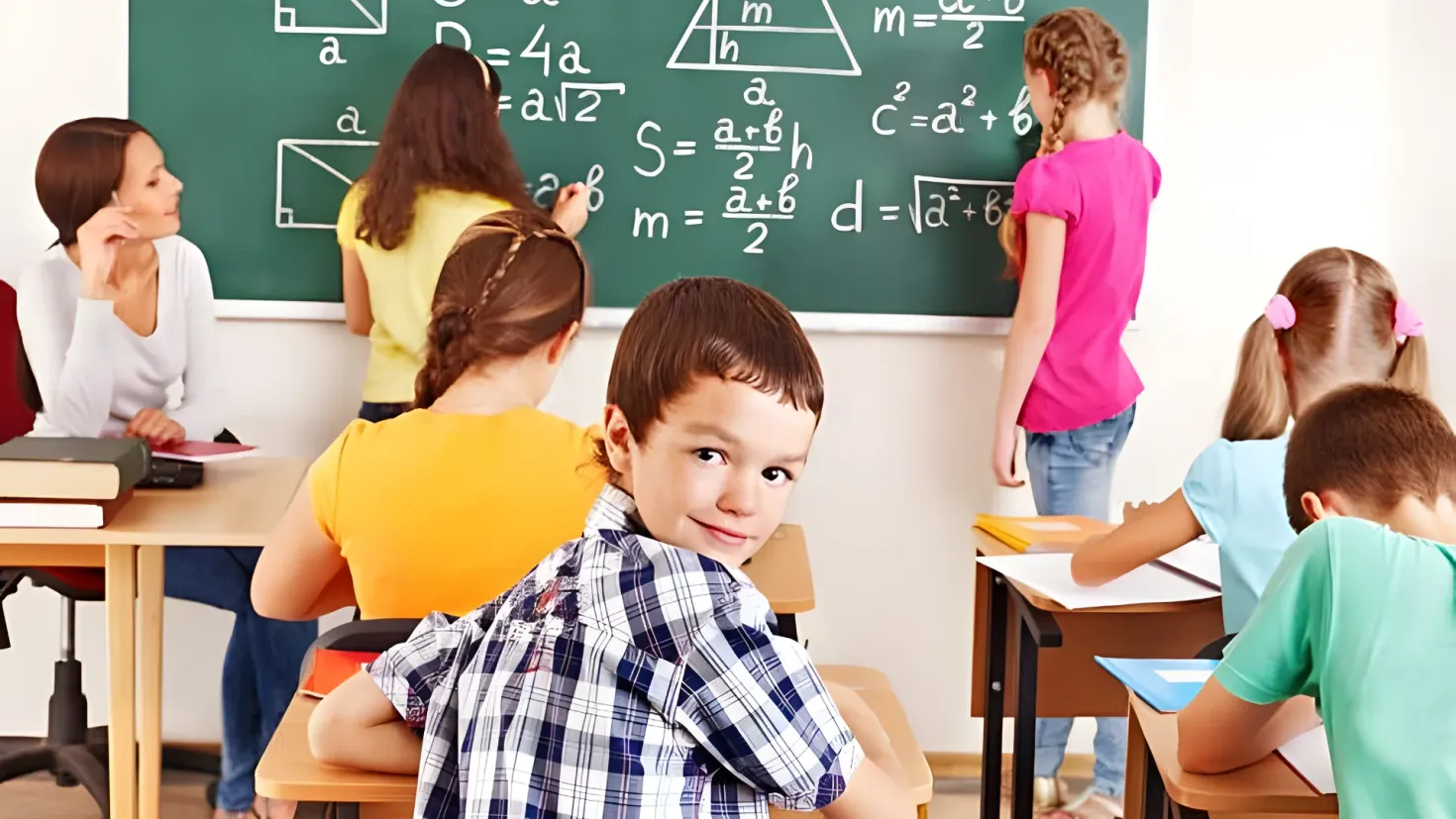 A group of children sitting at desks in front of a blackboard. There are books and other objects on the tables, and a woman standing on the floor. The blackboard is attached to the wall, and the children appear to be engaged in a math lesson.