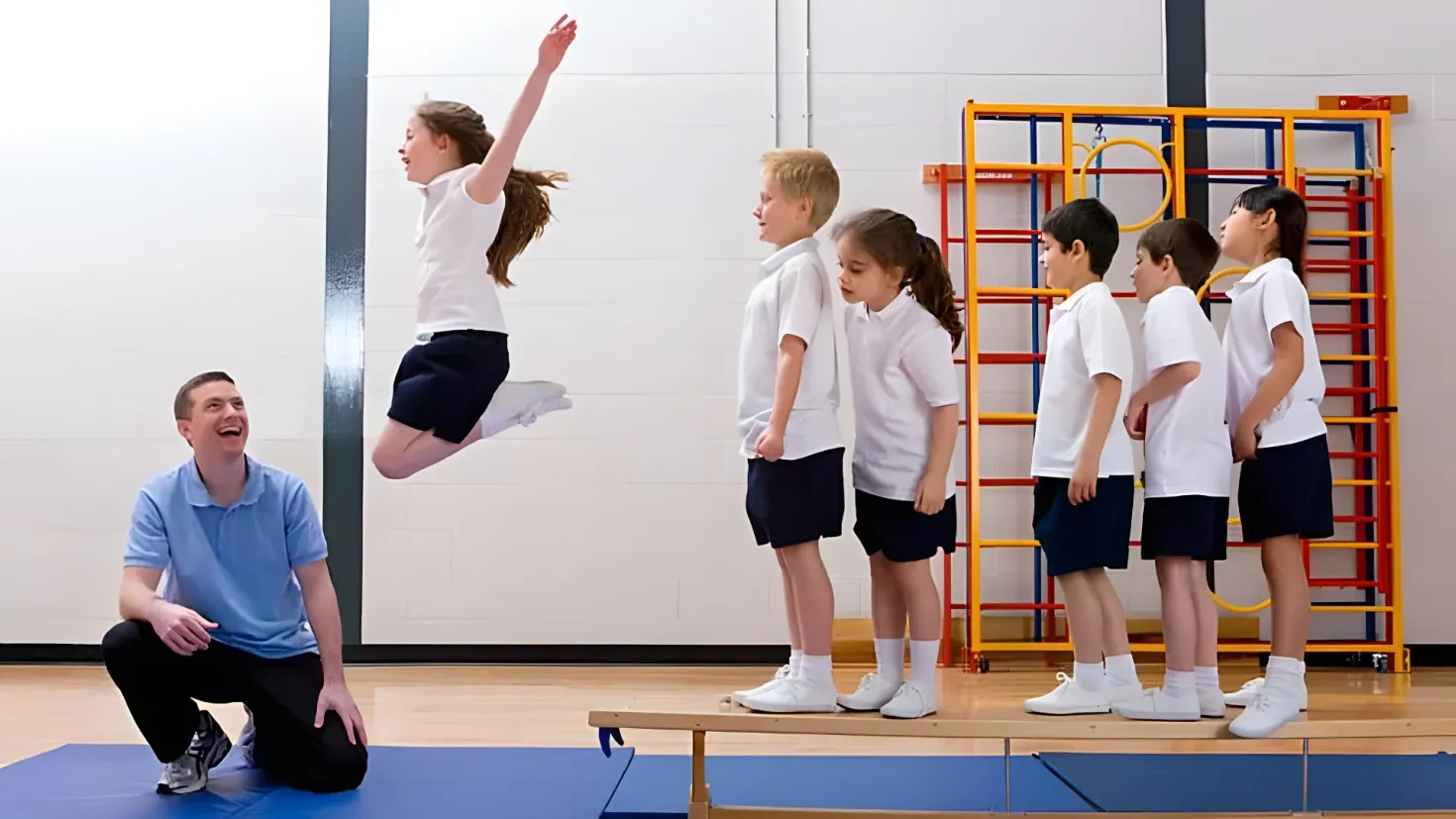 A group of children standing on top of a blue mat on a wooden floor, with a man in a squat position on the left side of the image. In the background, there is a wall and a ladder, suggesting that the children are in a gymnasium.