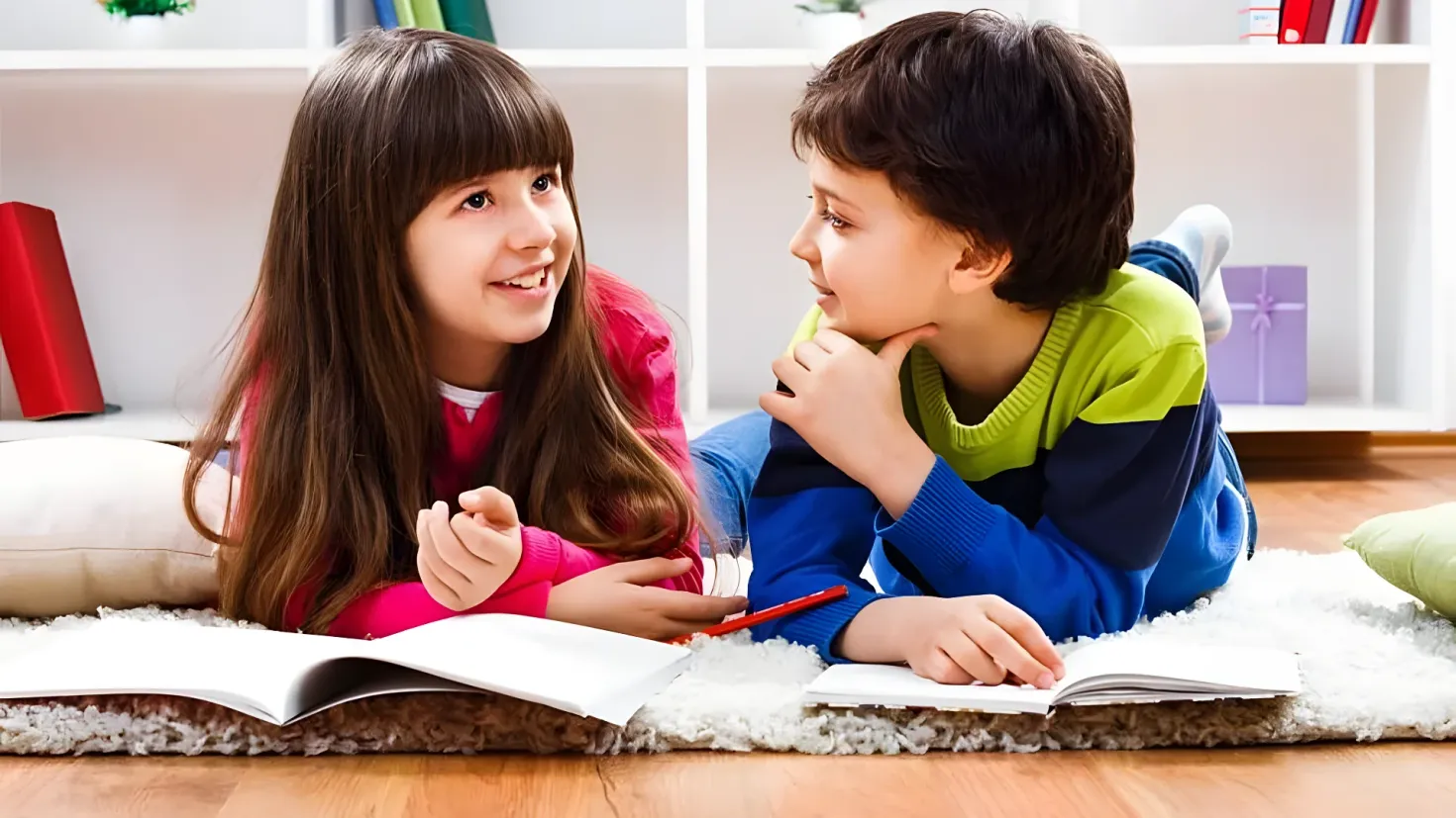 Two children, a boy and a girl, sitting on the floor and reading a book together. They are surrounded by pillows and a carpet, and in the background there are books and other objects in the racks.