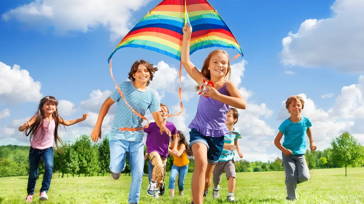 A group of children running across a lush green field, each of them holding a colorful kite in their hands. The sky is a bright blue with white fluffy clouds, and the sun is shining down on the children, creating a cheerful atmosphere.