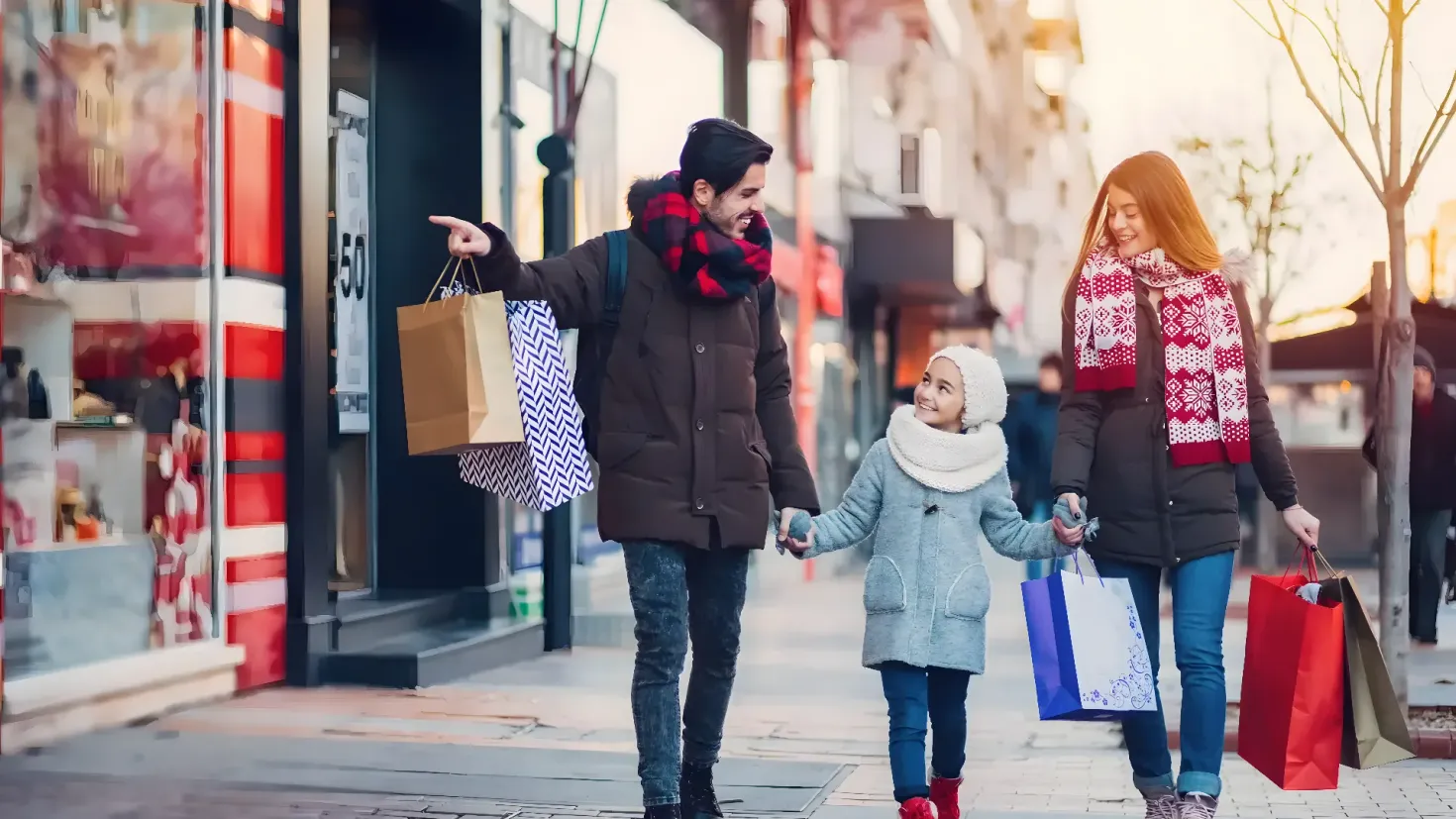 A family of three - a man, a woman and a child - walking down the street with shopping bags in their hands. The woman is wearing a cap and the child is holding a handbag. In the background, there are buildings, trees and a clear blue sky.