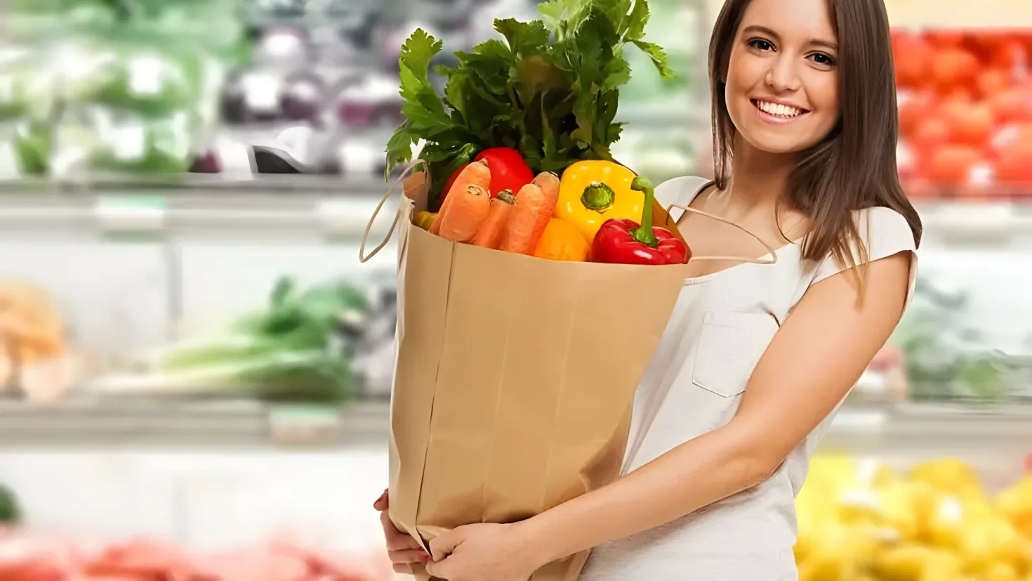 A woman standing and smiling while holding a paper bag full of vegetables in a grocery store. The background is slightly blurred, suggesting that the focus of the image is on the woman and the vegetables she is holding.
