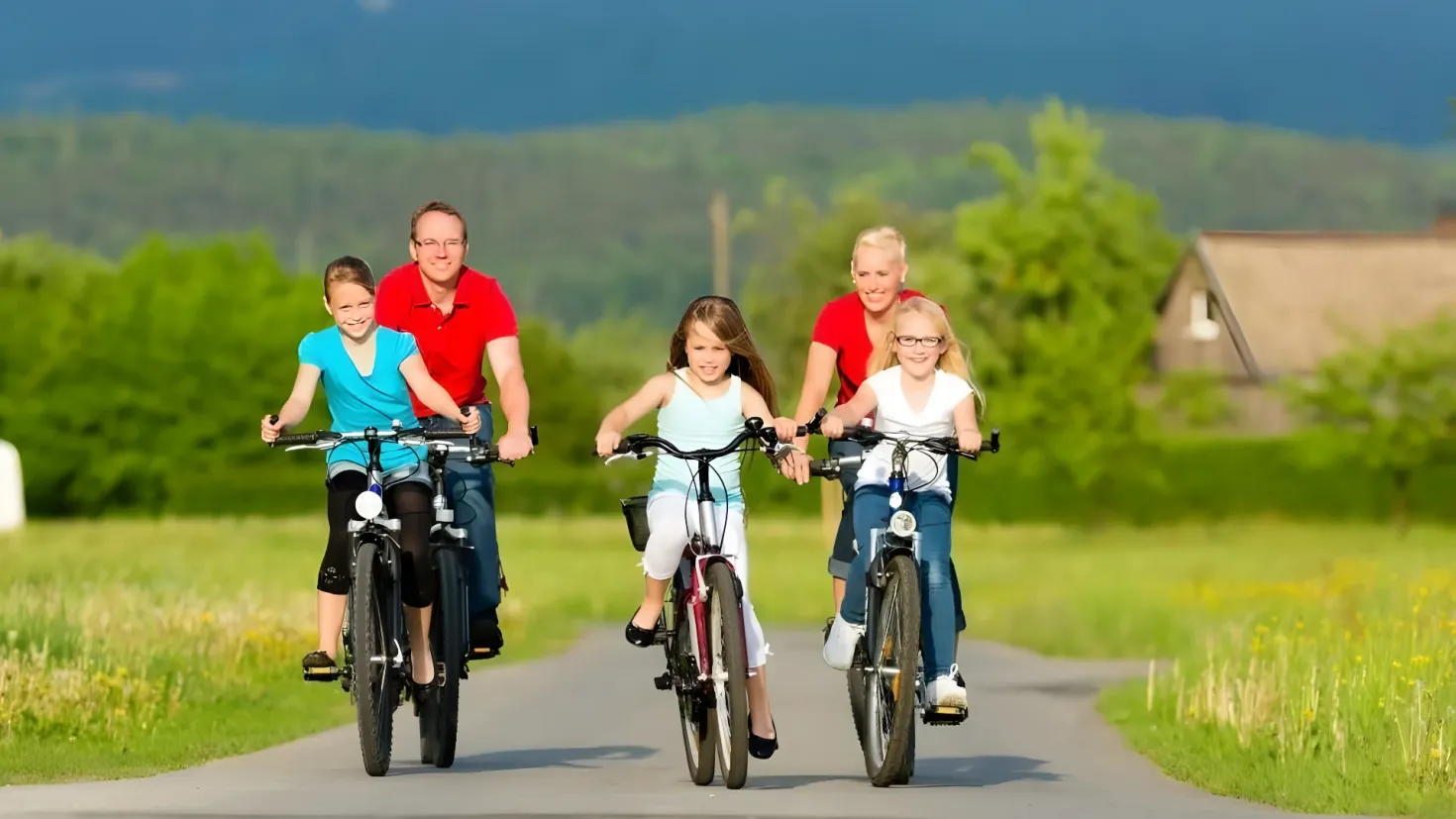 A family of four riding bicycles down a road surrounded by lush green grass, plants, and trees, with a house in the background and a bright blue sky above.